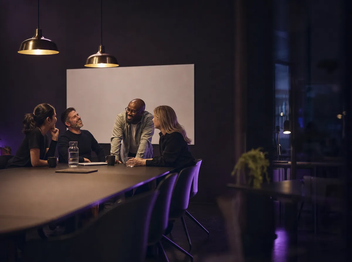 Four teammates talk around a table in a dark office, mid-conversation and relaxed.