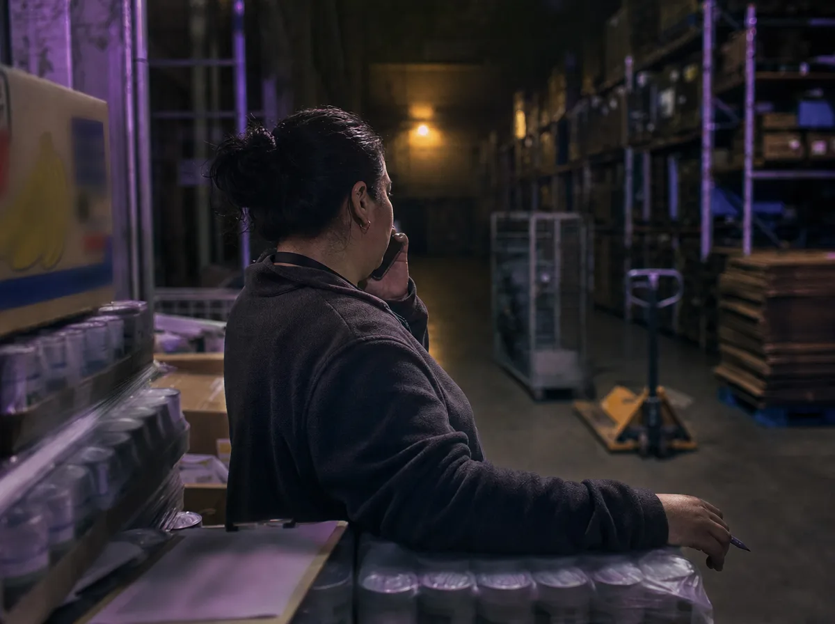 Food bank coordinator on a phone call leaning on a pallet of canned goods in a dark warehouse aisle.