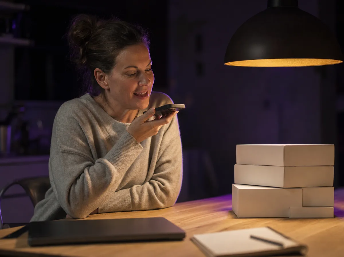A woman at her kitchen table at night records a message on her phone beside plain boxes.