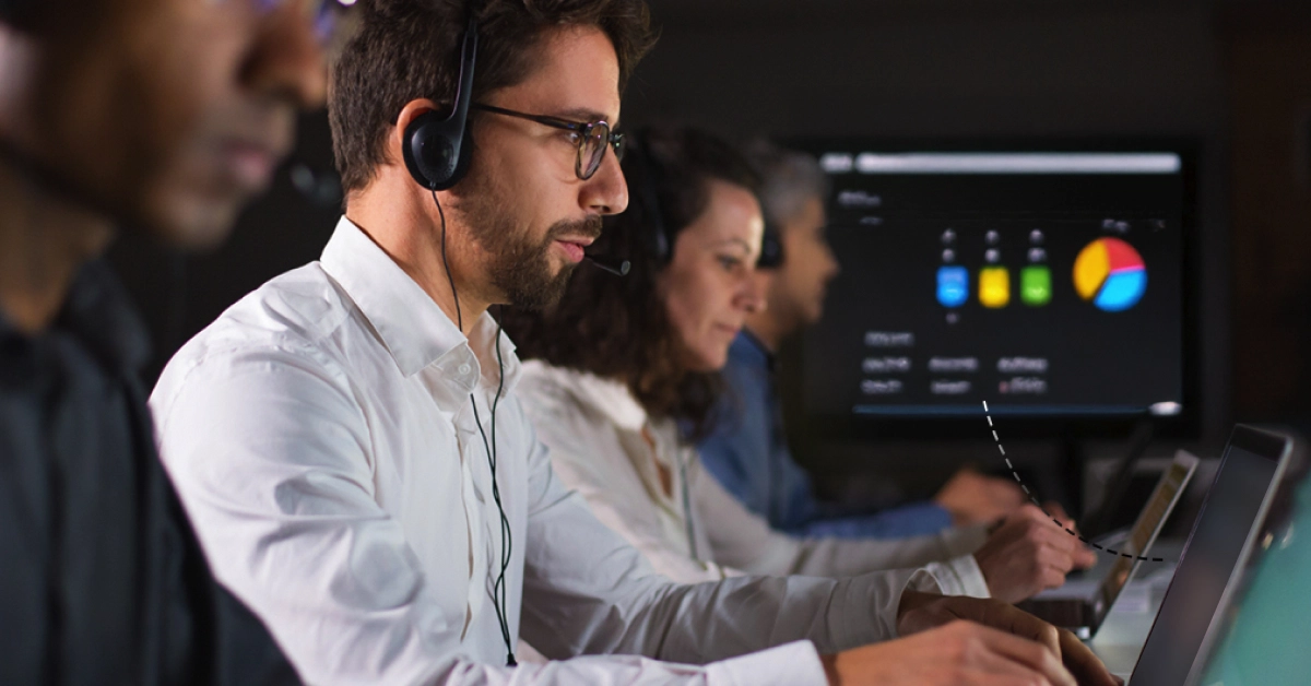 A group of four people sits in a row, wearing headsets and working on laptops, intensely focused on their screens. In the background, a large monitor displays colorful charts and data visualizations, likely analyzing AI call routing metrics to enhance performance. VoiceDrop Ringless Voicemails