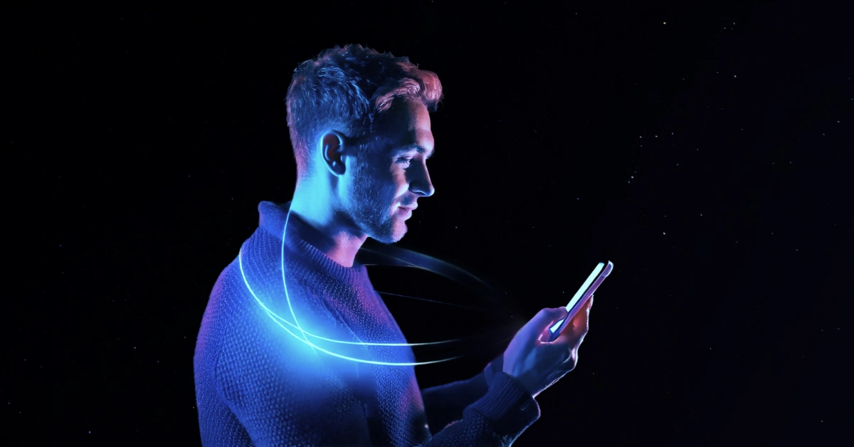 A man with a light beard, wearing a sweater, looks at his smartphone against a dark background. A glowing blue spiral encircles him, like AI-powered virtual agents in action. Small stars or dots are scattered in the background, enhancing the futuristic space-like theme. VoiceDrop Ringless Voicemails