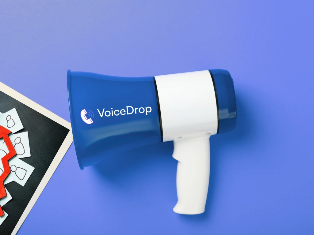 A blue and white megaphone labeled VoiceDrop lies on a purple surface next to a partial photo of red and white name tags with small badges, highlighting the event where Tim Bilyeu made a special appearance. VoiceDrop Ringless Voicemails