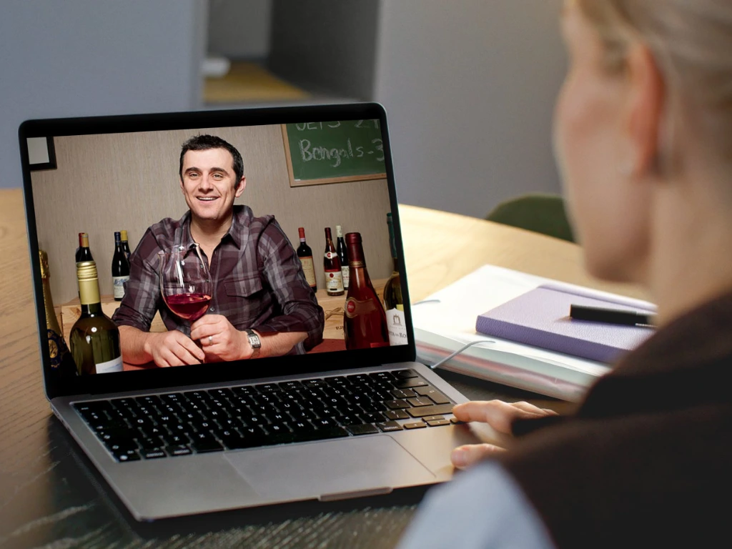 A person watches Gary Vaynerchuk holding a glass of red wine during a video call on a laptop. Several wine bottles are visible behind him on screen, while the viewer sits at a table with notebooks and papers. VoiceDrop Ringless Voicemails