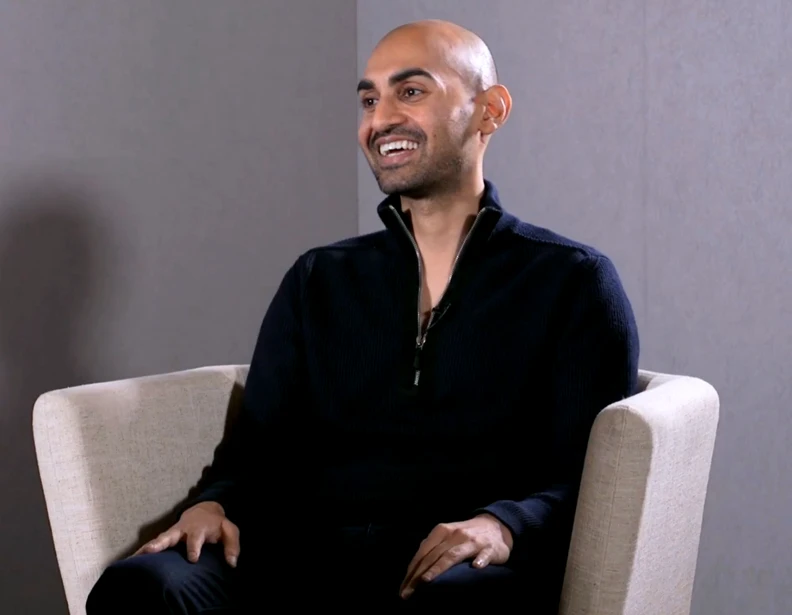 A man with a shaved head and beard, resembling Neil Patel, wearing a dark zip-up sweater, is sitting and smiling in a light-colored armchair against a plain, neutral background. VoiceDrop Ringless Voicemails