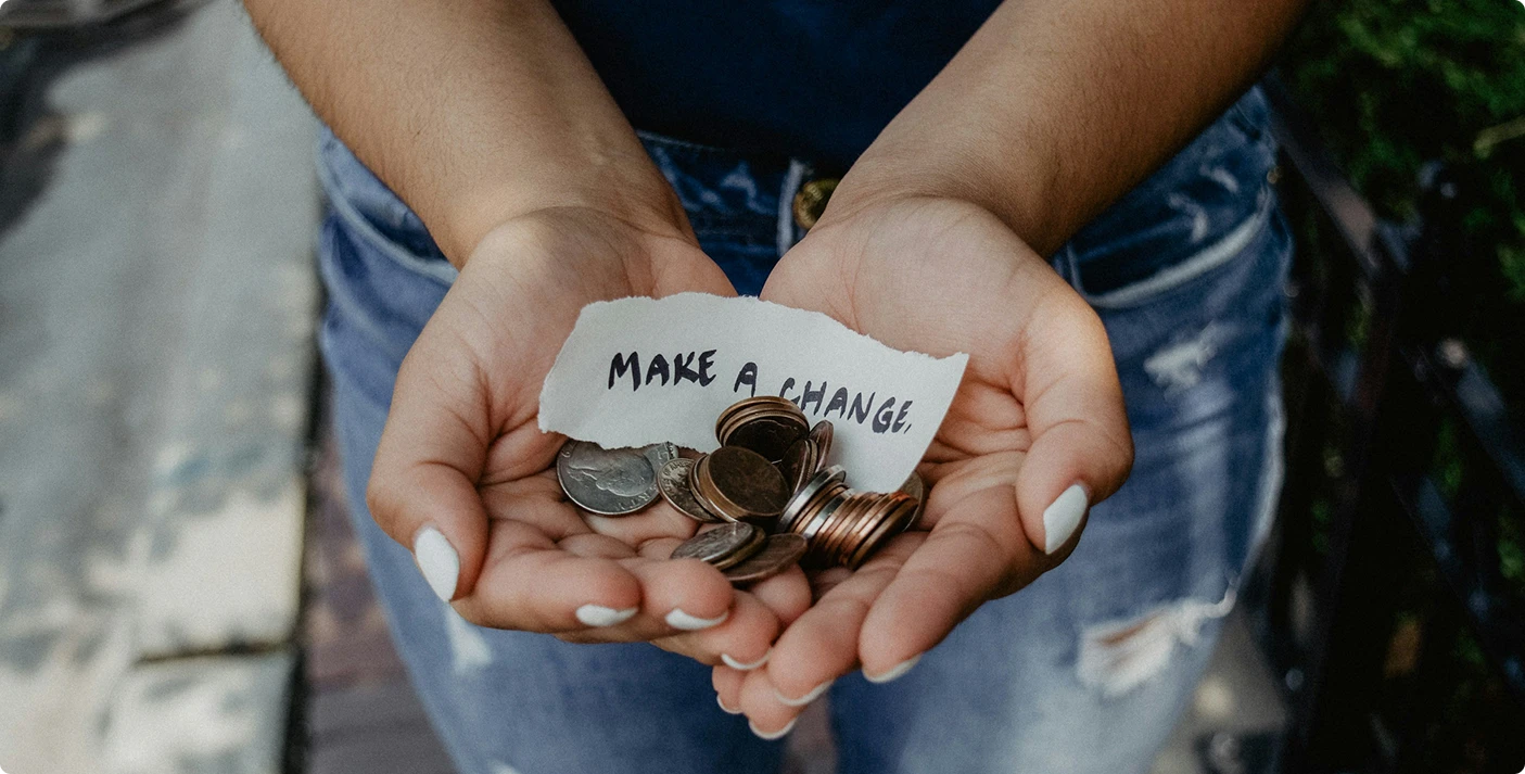 Close-up of hands holding various coins and a small torn piece of paper with the words Make a Change written on it, subtly suggesting support for nonprofit organizations. The background shows a blurred view of a person wearing jeans. VoiceDrop Ringless Voicemails