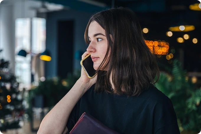 A woman with shoulder-length brown hair is holding a smartphone to her ear, looking thoughtful. She wears a black shirt and stands indoors, perhaps contemplating a career opportunity, with blurred lights and greenery in the background. VoiceDrop Ringless Voicemails