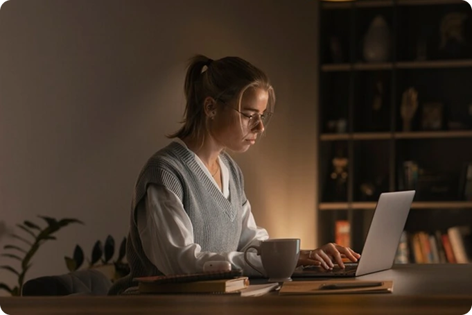 A career-driven individual with glasses and a ponytail works on a laptop at a desk in a dimly lit room. They wear a gray sweater vest over a white shirt. Books, a notebook, and a mug sit on the desk, with shelves and a plant completing the focused atmosphere. VoiceDrop Ringless Voicemails